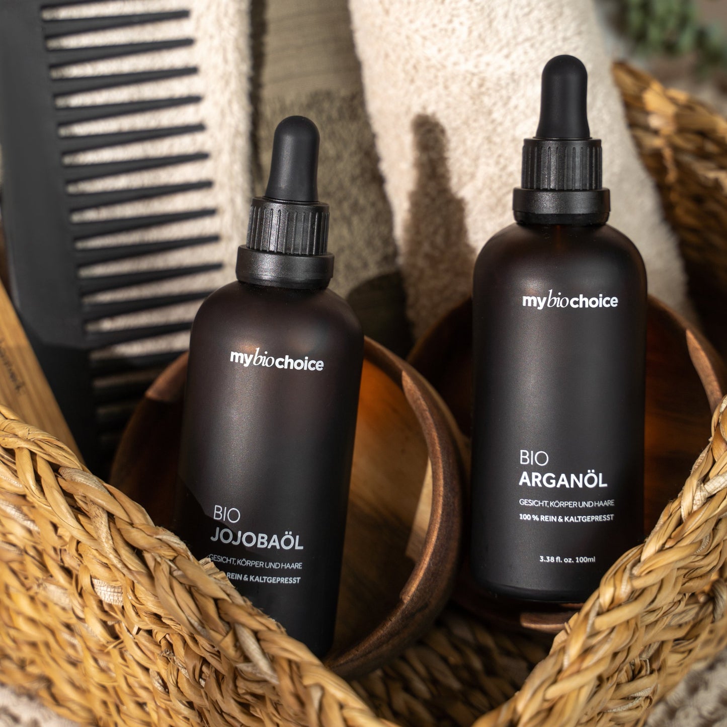 mybiochoice Bio Jojobaöl- und Bio Arganöl-Flaschen in einem Korb mit grünen und beigen Handtüchern und einem Kamm.