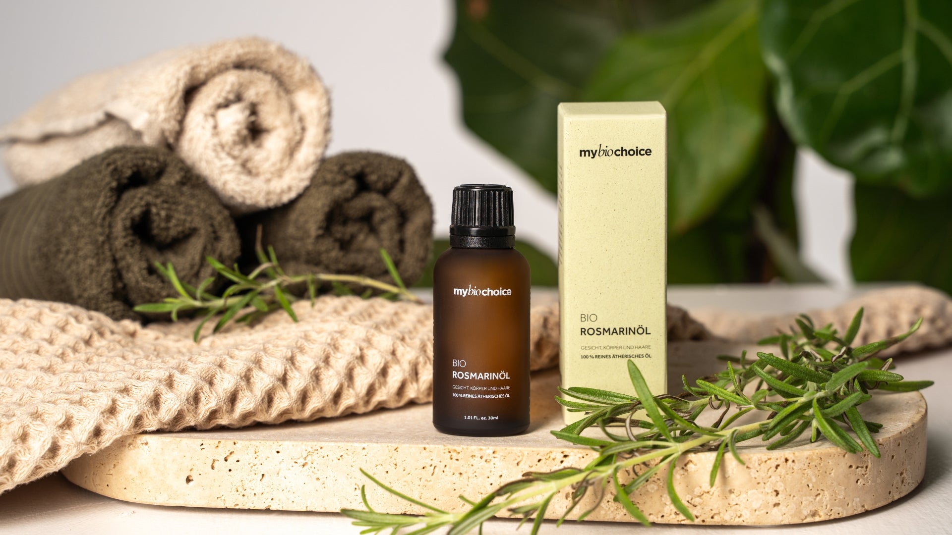 Bio Rosmarinöl von mybiochoice mit Verpackung, präsentiert auf Naturstein neben gerollten Handtüchern und Rosmarinzweig.