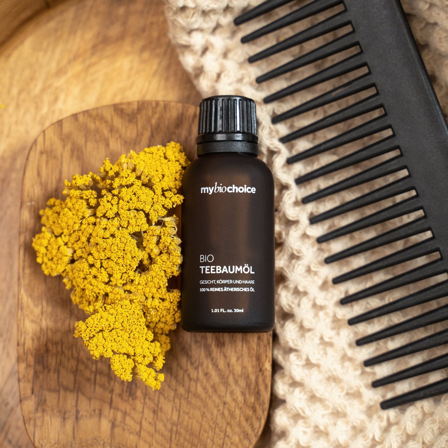 mybiochoice Bio Teebaumöl-Flasche mit gelben Blumen, einem Kamm und beigem Stoff, präsentiert in einem Holztablett.
