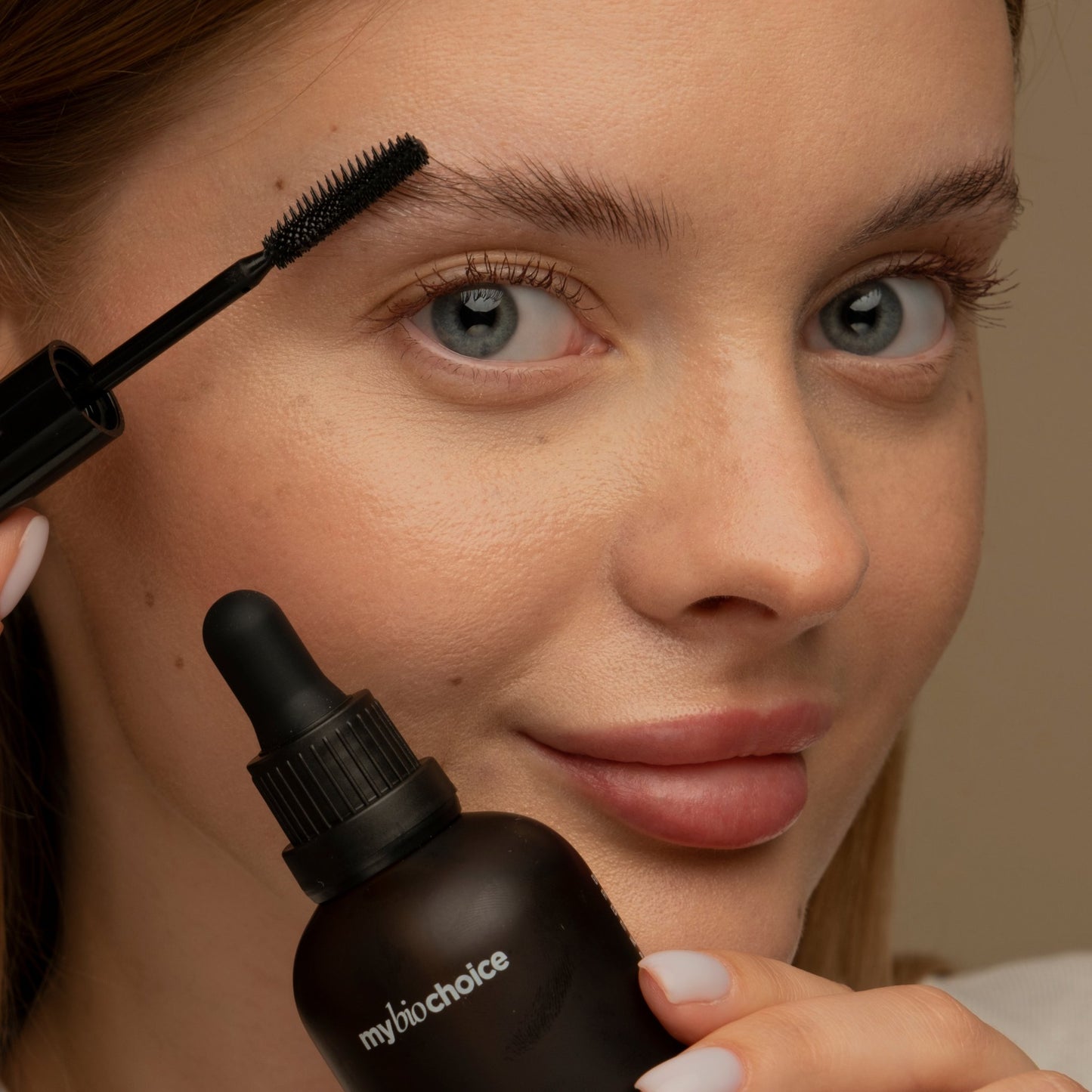 Eine Frau hält "mybiochoice" Bio Rizinusöl, bürstet ihre Augenbraue mit einer Mascara-Bürste, die Augen offen und lächelnd.