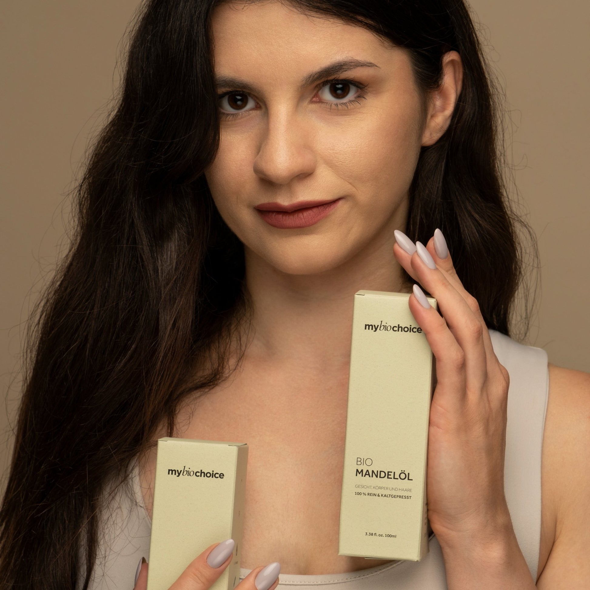 Eine Frau mit langem dunklem Haar hält zwei Schachteln Bio Mandelöl von "mybiochoice" und posiert in einem neutralen Oberteil.