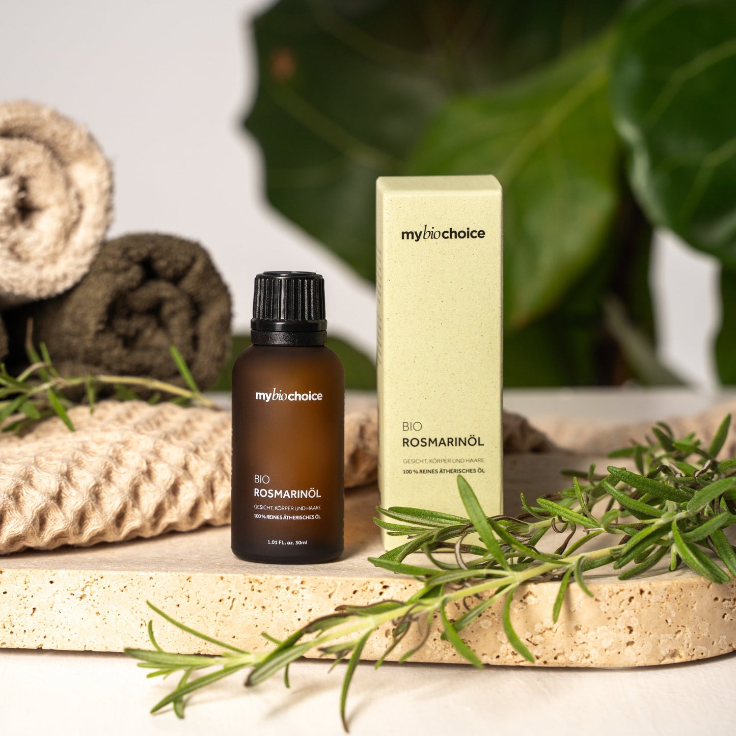 Mybiochoice Bio Rosmarinöl 30 ml mit grünem Karton auf einer Steinplatte, umgeben von Rosmarinzweigen und dekorativen Handtüchern, zur Pflege von Haut und Haar.