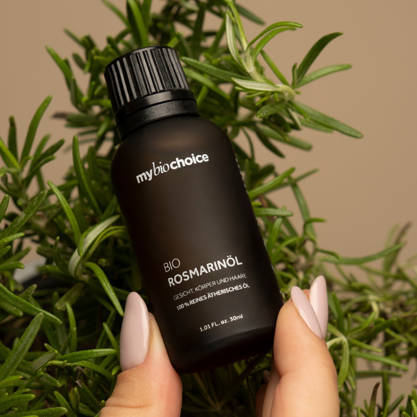Nahaufnahme einer Flasche Bio Rosmarinöl von "mybiochoice", gehalten vor Rosmarinzweigen.