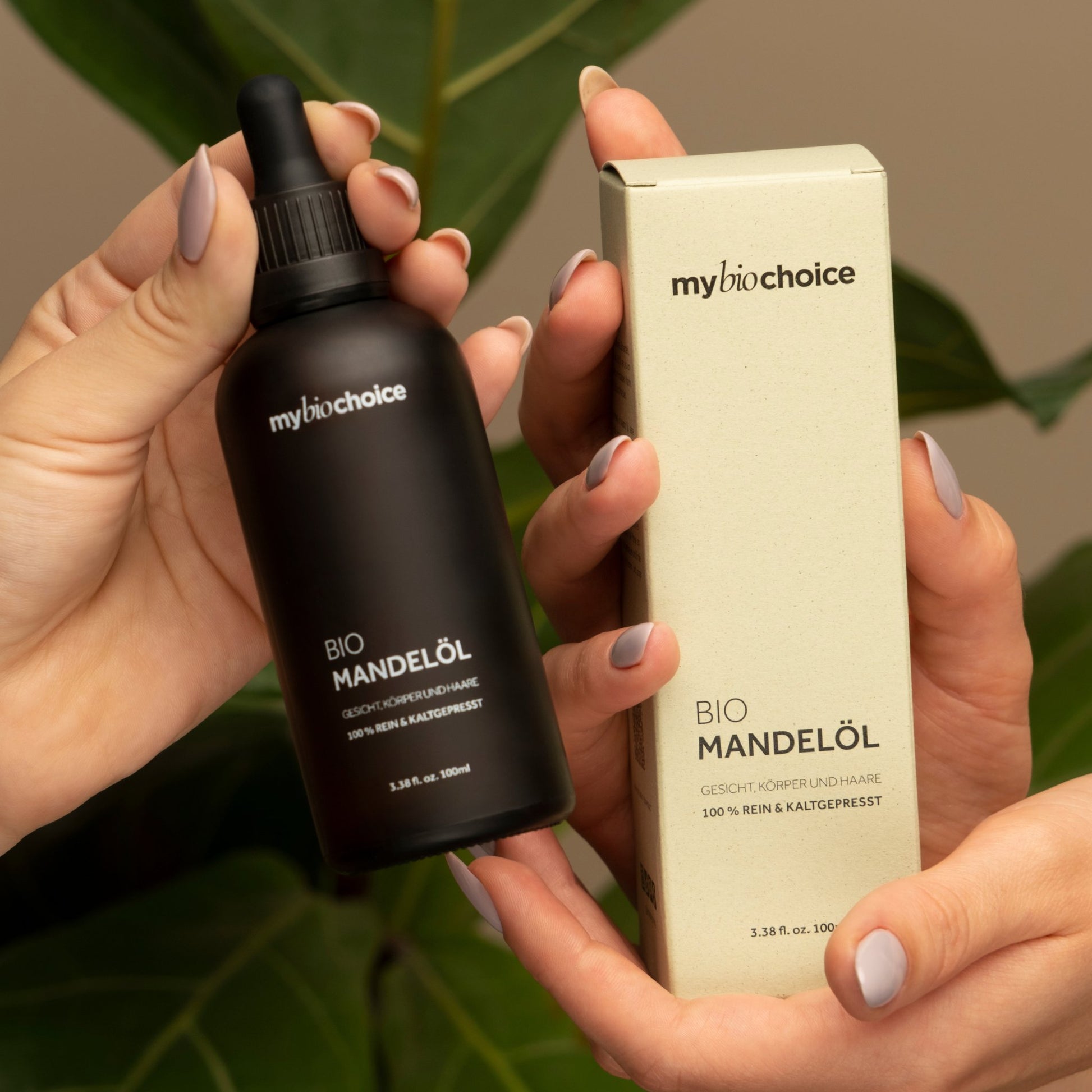 Nahaufnahme von Händen, die eine braune Flasche und eine Schachtel Bio Mandelöl von "mybiochoice" halten, mit grünen Blättern im Hintergrund.