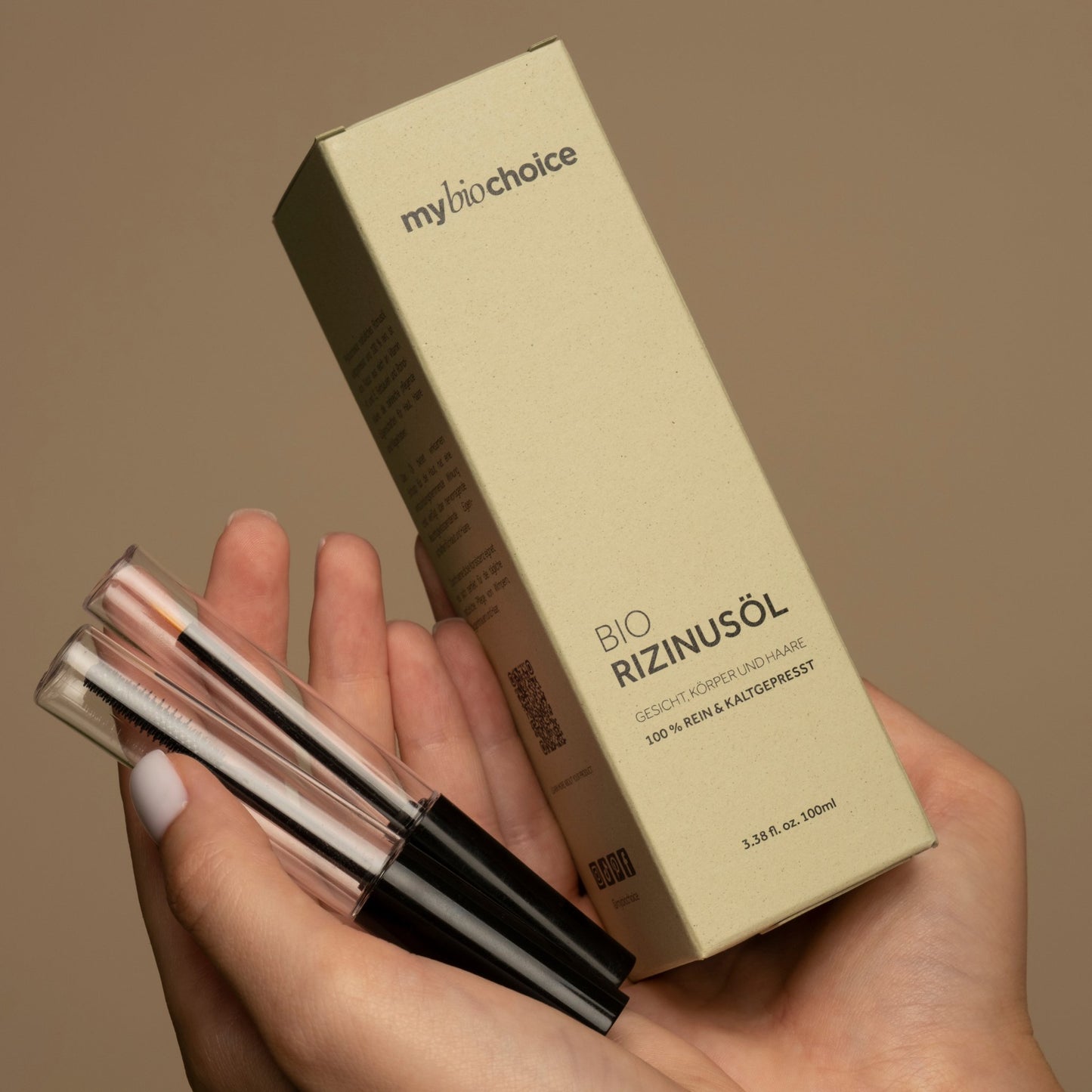 Nahaufnahme von Händen, die die Verpackung von "mybiochoice" Bio Rizinusöl und zwei Applikatoren für Augenbrauen- und Wimpernpflege halten.