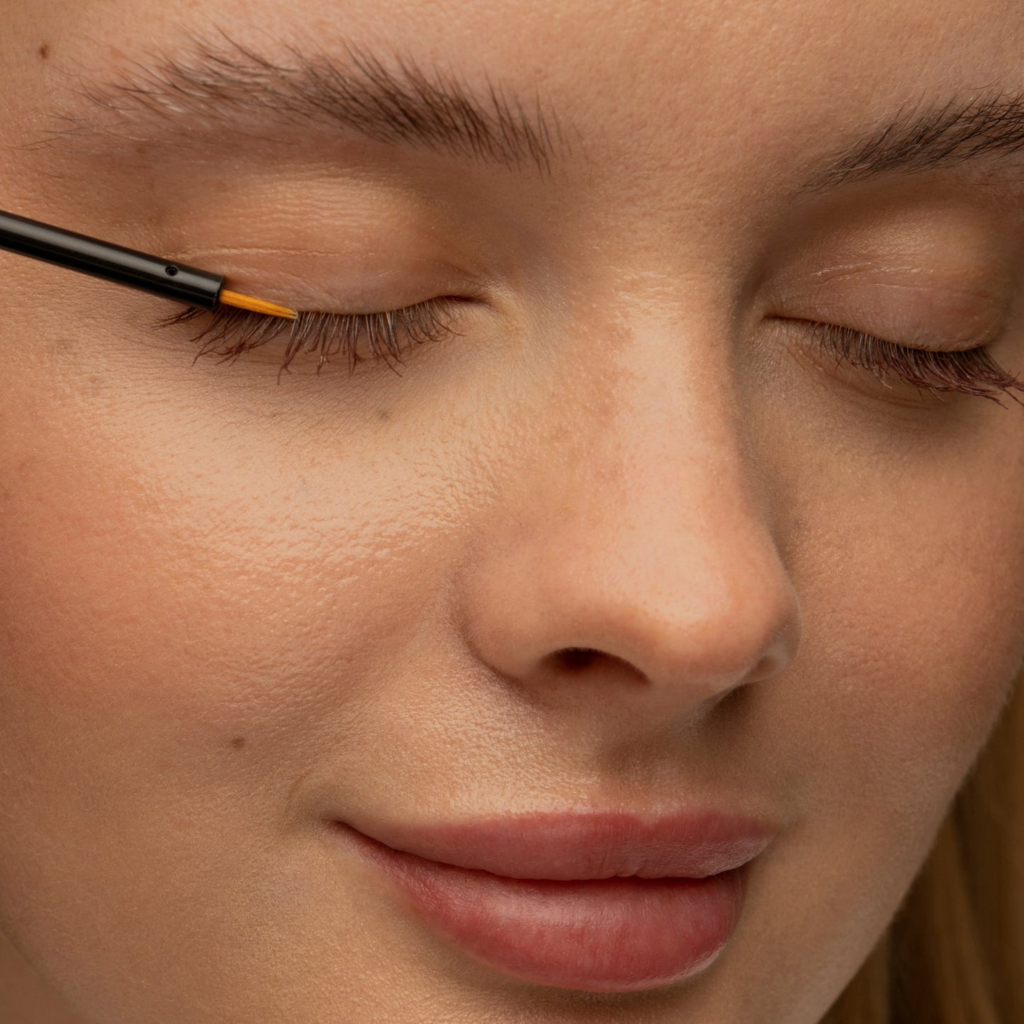 Nahaufnahme einer lächelnden Frau mit geschlossenen Augen, die "mybiochoice" Bio Rizinusöl mit einem dünnen Pinsel auf ihre Wimpern aufträgt.