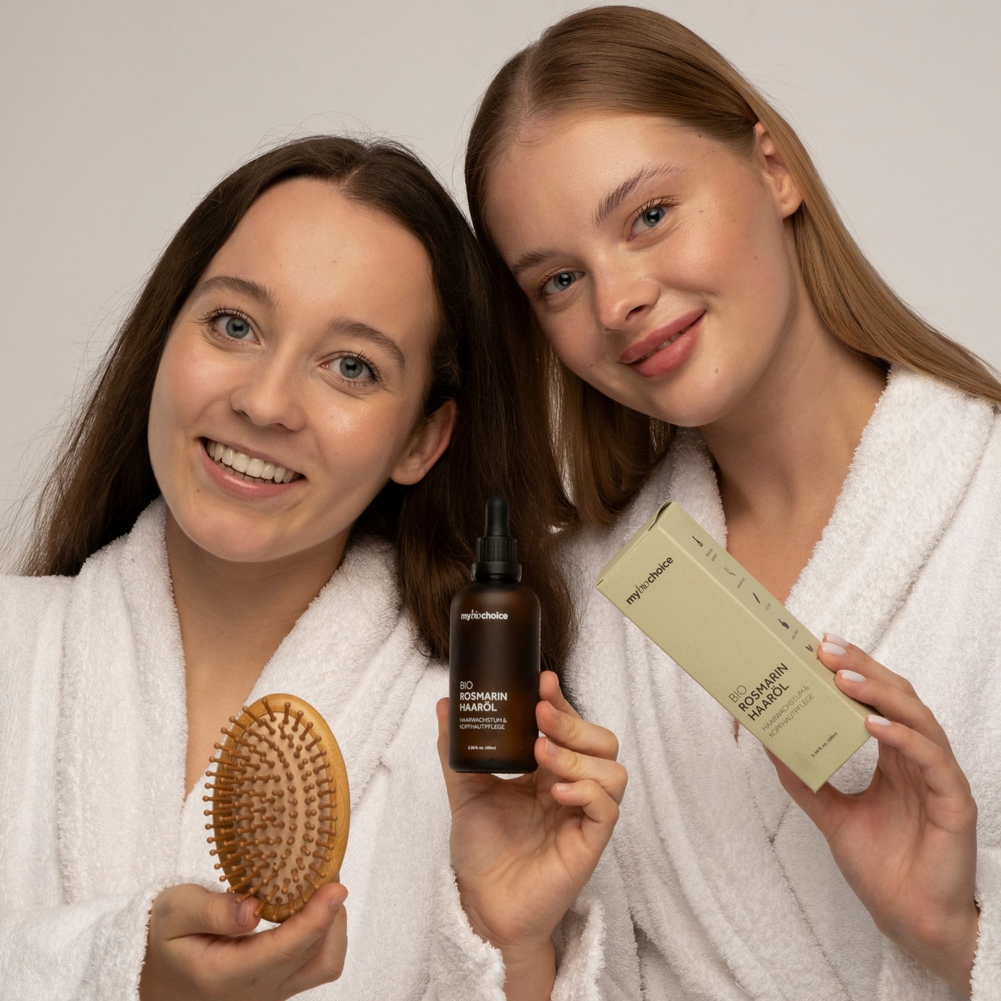 Zwei Frauen in weißen Roben lächeln und halten eine "mybiochoice"-Bambushaarbürste, eine Flasche Bio Rosmarin Haaröl und die Verpackung.