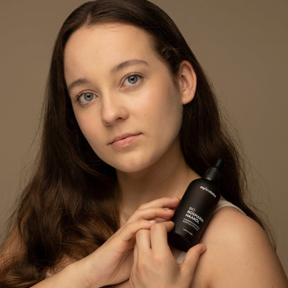 Eine Frau mit langem braunem Haar hält eine braune Flasche mit biologischem Rosmarin-Haaröl an ihre Schulter und posiert vor einem schlichten beigen Hintergrund.