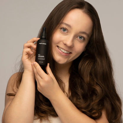 Eine Frau mit langem braunem Haar lächelt und hält eine Flasche "mybiochoice" Bio Arganöl.